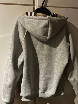 Grå zip hoodie från Burberry - Snygg grå zip hoodie från Burberry med klassiskt rutigt foder i huvan och dragsko. Hoodien har två fickor framtill och ribbade muddar vid ärmar och nederkant. Perfekt för en chill och stilren look. Är L men passar mer som M fråga gärna om ni har funderingar 