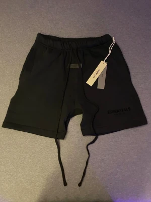 Essentials - Fear of God (Shorts) - Svarta shorts från Essentials Fear of God med snörning i midjan och diskret logga på benet. Mjukt material och avslappnad passform, perfekta för chill dagar. Klassisk design med fickor på sidorna och elastisk midja.