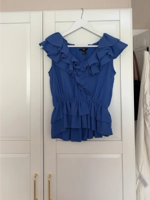Blå volangblus med v-ringning - Supersöt blå blus från H&M med volanger längs v-ringningen och midjan. 