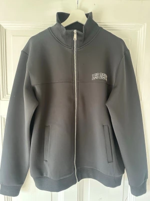 Svart zip hoodie från Les Deux - Snygg svart hoodie från Les Deux med hel dragkedja framtill och två fickor. Medel size design med Les Deux-logga på bröstet. Tillverkad i mjukt material och har hög krage. Perfekt för en clean och avslappnad look.