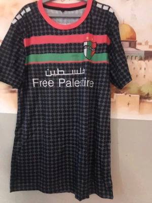 Palestina fotbollströja ny / xxxl - Snygg fotbollströja från Palestino med svart bas och prickigt mönster, röda och gröna ränder över bröstet samt texten 'Free Palestine'. Röd krage och emblem på bröstet. Baksidan har vit karta och text på arabiska och spanska. Perfekt för match eller träning.