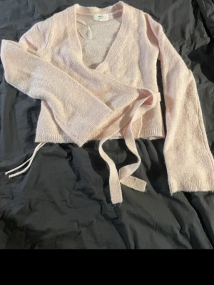Ljusrosa omlottkofta från Gina Tricot - Mysig ljusrosa stickad omlottkofta från Gina Tricot i ullblandning. Koftan har långa ärmar, djup v-ringning och knyts i midjan med band. Perfekt för lager-på-lager och en söt touch till din outfit. Den ser mer rosa ut I verkligheten!💕