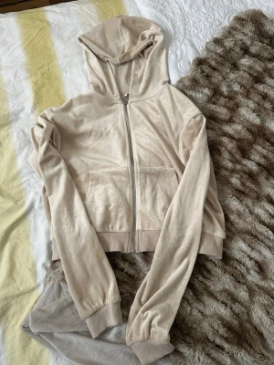 Beige zip-hoodie i mjuk velour - Säljer en beige hoodie i mjuk velour med dragkedja framtill och känguruficka. Modellen har ribbade muddar vid ärmslut och nederkant samt en klassisk huva. Perfekt för en avslappnad och trendig look.