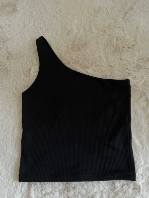 Svart oneshoulder topp Gina Tricot - Snygg svart oneshoulder topp från Gina Tricot i ribbad material. Toppen är tight och har en  asymmetrisk design med bred axelrem på ena sidan. Perfekt till både fest eller vardagstopp storlek xs 