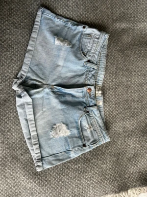 Ljusa denimshorts med slitningar - Säljer ett par ljusblå denimshorts med slitna detaljer framtill och klassiska fem fickor. Shortsen har uppvikta benslut och knappgylf. Perfekta för sommaren och ger en avslappnad vibe.
