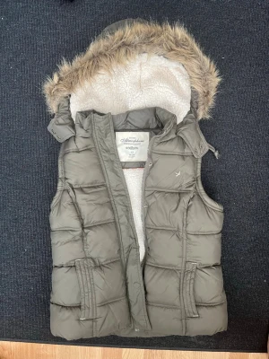 Olivgrön dunväst med fuskpälsluva - Snygg olivgrön dunväst från Atmosphere med avtagbar luva kantad med fuskpäls och mjukt vitt teddyfoder. Västen har två fickor framtill och stängs med dragkedja och tryckknappar. Perfekt för lager på lager och håller dig varm under kyliga dagar.