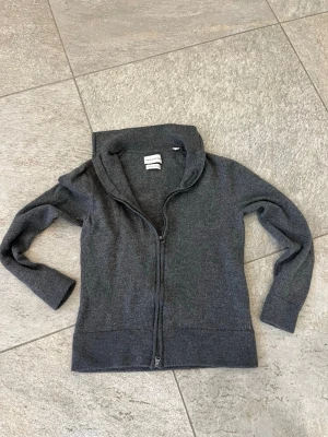 Mörkgrå ziptröja från Soft Goat - Mysig mörkgrå ziptröja från Soft Goat i mjuk ullblandning. Tröjan har hög krage, dragkedja framtill och ribbade muddar vid ärmslut och nederkant. Perfekt för lager på lager och enkel att matcha med olika outfits.