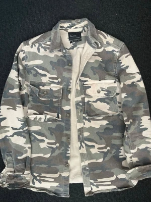 Camo overshirt från Zara - Säljer en camouflagemönstrad overshirt från Zara i relaxed fit. Skjortan har två stora bröstfickor med lock och knappar, klassisk krage och tryckknappar framtill. Materialet är kraftigt bomullstyg i grå, beige och gröna toner.