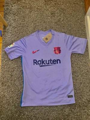 Lila FC Barcelona matchtröja Nike - Snygg lila FC Barcelona matchtröja från Nike med Dri-FIT-material. Tröjan har V-ringning, klubbmärke och sponsorlogga på bröstet samt La Liga-märke på ärmen. Perfekt för dig som älskar fotboll och vill representera Barca.