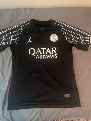 Svart PSG x Jordan matchtröja - Snygg svart Paris Saint-Germain matchtröja från Jordan med vita detaljer och loggor. Tröjan har tryck av Qatar Airways på bröstet och coola vita mönster på ärmarna. Tillverkad i lätt och ventilerande Dri-FIT-material, perfekt för fotboll eller träning.