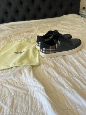 Burberry sneakers i marinblått - Snygga marinblå sneakers från Burberry med klassiskt rutigt mönster på sidorna och vit sula. Skorna har lågt skaft och är tillverkade i en mix av läder och textil. Perfekta för dig som gillar stilrena och exklusiva detaljer.