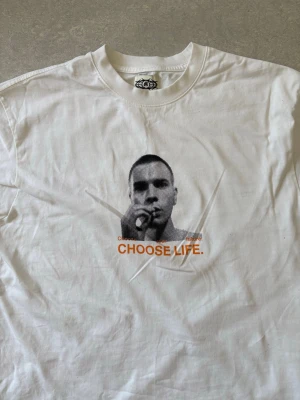 Vit t-shirt med Choose life motiv box fit - Vit t-shirt från Zoo York med stort tryck av ansikte och texten 'CHOOSE LIFE.' i orange framtill. Baksidan har ett långt texttryck och samma slogan i svart och orange. Klassisk passform och rund hals. Perfekt för dig som gillar statement-plagg. Baserat på filmen trainspotting 