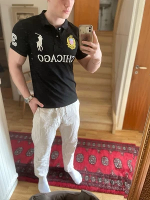 Svart Ralph Lauren piké Chicago - Svart piké från Ralph Lauren med klassisk pikékrage, stora vita tryck på bröstet och ryggen samt broderad logga och klubbmärke. Kortärmad modell i bomull med sportiga nummerdetaljer på ärm och rygg.