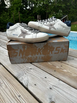 Golden Goose vita sneakers med stjärna - Golden Goose sneakers i vitt läder med grå mockadetaljer och ikonisk stjärna på sidan. Skorna har mörkblå hälkappa, grå snören och slitna detaljer för en cool vintage-look. Perfekta för dig som gillar streetstyle och unika detaljer.