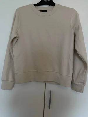 Beige sweatshirt från ZARA - En stilren beige sweatshirt från ZARA med rund halsringning och ribbade muddar vid ärmslut och nederkant. Tröjan har en avslappnad passform och är tillverkad i mjukt bomullsmaterial, perfekt för en clean och enkel look. Storlek S. Andra bilden är inte på mig utan exempel bild hur de hade suttit på. Använt en gång så ser helt ny ut. 