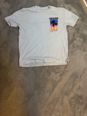 Vit t-shirt med Miami-tryck - Vit t-shirt med färgglatt Miami-tryck på både fram- och baksidan. Motivet visar palmer mot en solnedgång i lila, rosa och orange toner samt texten 'MIAMI Florida'. Perfekt för dig som gillar retro och streetwear vibes.
