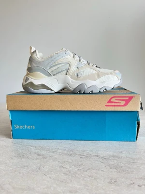  Sneakers från Skechers - Chunky sneakers från Skechers i vitt och ljusgrått med detaljer i mesh och syntet. Tjock sula med vågig design och klassisk snörning. Perfekta för dig som gillar sportig och trendig stil.