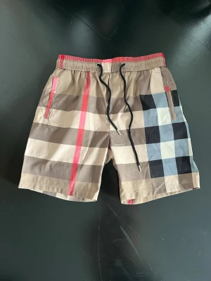 Rutig Burberry bad shorts - Säljer ett par klassiska Burberry shorts med ikoniskt rutigt mönster i beige, svart, rött och vitt. Shortsen har elastisk midja med svart snörning och fickor på sidorna även baken. Perfekta för sommaren och riktigt snygga till streetwear-stilen.