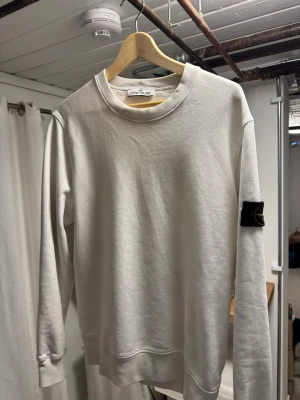 Vit sweatshirt från Stone Island - Vit sweatshirt från Stone Island med klassisk rund halsringning och ikonisk svart patch med logga på vänster ärm. Tröjan har en avslappnad passform och är tillverkad i mjuk bomull. Perfekt för en clean och stilren look. Nästan aldrig använd  max 2-3 gånger varit för stror det är Storlek M men kan passa L