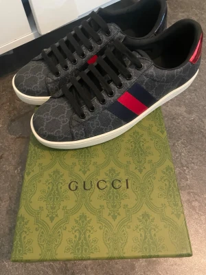 Gucci ace - Snygga svarta Gucci sneakers med klassiskt GG-mönster i grått, röd och blå rand på sidan samt vit sula. Skorna har svarta skosnören och en röd detalj på hälen. Tillverkade i canvas och läder för en exklusiv känsla.