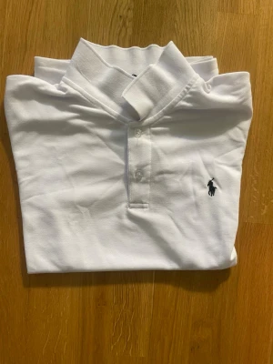 Vit piké från Polo Ralph Lauren - Klassisk vit pikétröja från Polo Ralph Lauren med broderad logga på bröstet. Skjortan har krage, knappar framtill och är tillverkad i bomull. Perfekt för en clean och stilren look.