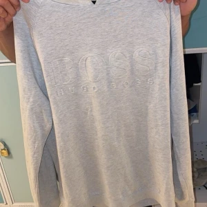 Ljusgrå hoodie från Hugo Boss - Snygg ljusgrå hoodie från Hugo Boss med diskret broderad logga på bröstet. Hoodien har långa ärmar, rundad halsringning och är gjord i mjukt bomullsmaterial. Perfekt för en clean och stilren look.