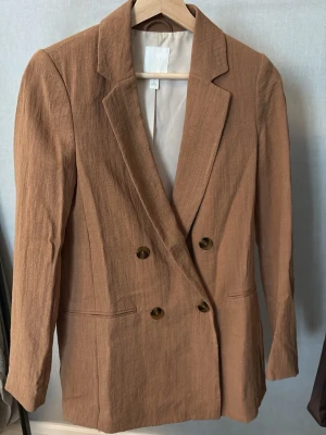 Beige dubbelknäppt kavaj H&M - Stilren beige kavaj från H&M med dubbelknäppning och klassisk krage. Kavajen har två framfickor och är tillverkad i ett lätt, strukturerat tyg som ger en avslappnad men ändå dressad look. Perfekt för att lyfta vilken outfit som helst.
