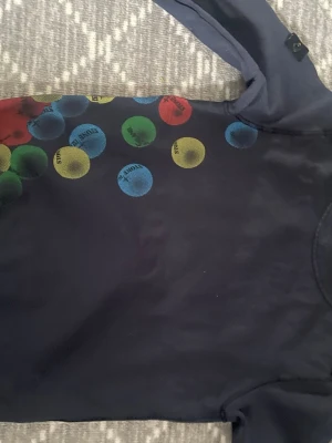 Mörkblå långärmad t-shirt Stone Island - Mörkblå långärmad t-shirt från Stone Island med färgglada prickar i rött, blått, grönt och gult på axel och nederkant. Har en liten ficka på bröstet och klassisk Stone Island-patch på ärmen. Tillverkad i mjuk bomull.