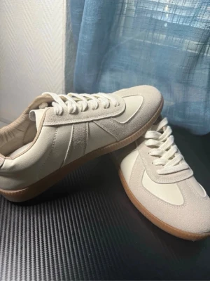 Maison margiela sneakers - Snygga beige sneakers med ovandel i läder och mockadetaljer. Skorna har klassisk låg profil, vita snören och en brun gummisula som ger en retrovibe. Perfekta för dig som gillar stilrena och tidlösa sneakers. Ej äkta (ÖPPEN FÖR BYTEN OCKSÅ)