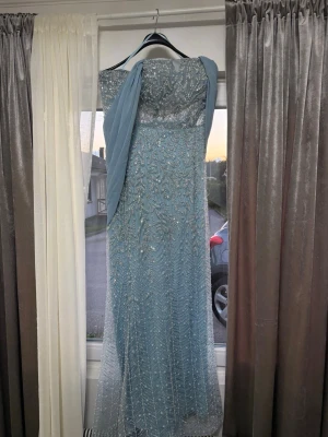 Ljusblå glittrig långklänning - Elegant ljusblå långklänning med glittriga paljettdetaljer och broderier. Klänningen har en vacker draperad sjal över ena axeln och öppen rygg. Perfekt för bal eller festliga tillfällen. Materialet känns som polyester eller syntet. Den är använd endast 1 gång för bröllop. Nypris:2600