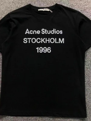 Svart Acne Studios t-shirt - Svart t-shirt från Acne Studios med vit text 'Acne Studios STOCKHOLM 1996' på bröstet. Klassisk rund hals och korta ärmar. Perfekt för en stilren och trendig look.