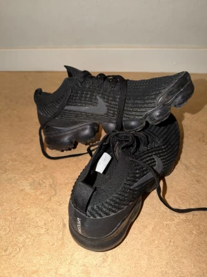 Nike Vapormax svarta sneakers - Säljer ett par helsvarta Nike Vapormax sneakers med stickad ovandel och synlig logga på sidan. Skorna har en unik, bubblig sula i gummi och texten 'VAPORMAX' på hälen. Perfekta för dig som gillar sportig och modern stil.