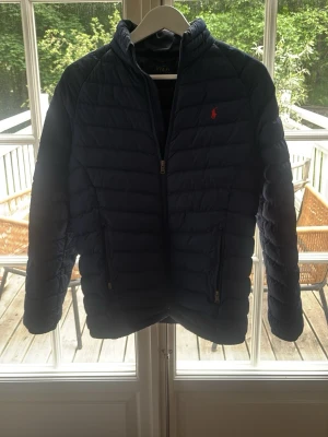 Mörkblå pufferjacka från Polo Ralph Lauren - Snygg mörkblå pufferjacka från Polo Ralph Lauren med klassisk röd logga på bröstet. Jackan har dragkedja framtill, två sidofickor med dragkedja och quiltad design. Perfekt för kalla dagar och ger en sportig look.
