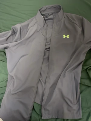 Grå träningsjacka från Under Armour - Snygg grå träningsjacka från Under Armour med dragkedja och hög krage. Jackan har ett neongult logotryck på bröstet och stort UNDER ARMOUR-tryck på ryggen. Tillverkad i ett mjukt och stretchigt funktionsmaterial som passar perfekt till träning.