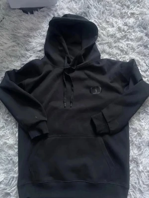 Svart hoodie  - Säljer en svart hoodie med broderad logga på bröstet. Hoodien har klassisk känguruficka, dragsko i huvan och ribbade muddar vid ärmslut och nederkant. Perfekt för en chill och avslappnad stil.
