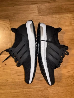 Svarta Adidas Ultraboost sneakers - Säljer ett par svarta Adidas Ultraboost sneakers med klassiska tre ränder på sidan. Ovandelen är i mesh och syntet, sulan är vit med svart yttersula i Continental-gummi. Skorna har en sportig och clean look, perfekta för dig som gillar streetwear.