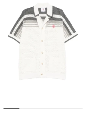 Stickat set från Casablanca i vitt och grått - Säljer ett stickat set från Casablanca med skjorta och shorts i vitt och grått samt vitt och blått. Skjortan har kort ärm, knappar framtill, två fickor och en röd logga på bröstet. Shortsen har dragsko i midjan, gråa ränder nertill och matchande röd logga. Materialet är virkat och känns luftigt.