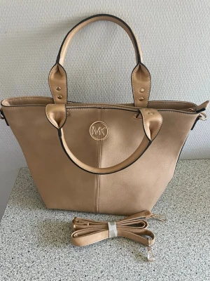 Beige handväska MK - Stilren beige handväska i skinnimitation med guldfärgade detaljer och MK-logga framtill. Väskan har både handtag och avtagbar axelrem.