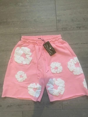 Demin tears shorts rosa - Säljer ett par rosa shorts från Denim Tears med vita blommiga applikationer. Shortsen har resårmidja med snörning och avslappnad passform. Perfekta för sommaren och riktigt snygga till en enkel t-shirt.