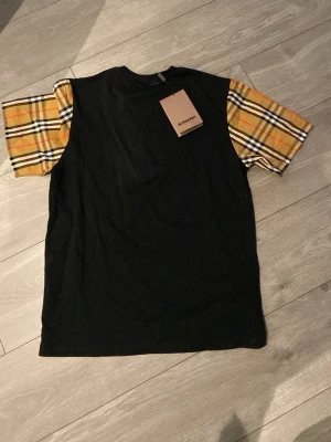 Burberry t-shirt med rutiga ärmar - Snygg svart t-shirt från Burberry med klassiska gul-bruna rutiga kortärmar. T-shirten har en rak passform och är tillverkad i mjuk bomull. Perfekt för dig som vill ha en unik twist på din outfit! Storlek M men funkar bra som L