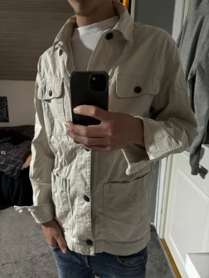 Beige utilityjacka - Stilren beige utilityjacka med fyra framfickor och svarta knappar. Jackan har en rak passform och är tillverkad i ett lätt bomullsmaterial, perfekt för lager på lager. Långärmad och enkel att matcha till olika outfits.