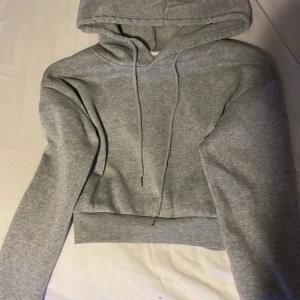 Grå croppad hoodie med huva - Snygg grå croppad hoodie med huva och dragsnören. Tillverkad i mjukt sweatshirtmaterial och har bred mudd i midjan samt långa ärmar. Perfekt för en avslappnad och trendig look.