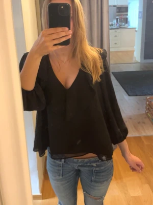 Svart blus med volang från Zara - Svart blus från Zara med v-ringning och volangdetalj längs halsringningen. Blusen har långa, lite puffiga ärmar med resår vid ärmslut och är tillverkad i ett lätt, flowy tyg som ger en snygg siluett. Man kan både klä upp och klä ner tröjan💕