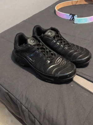 Nike Air Max Plus helsvarta sneakers - Nike Air Max Plus sneakers i helsvart med mesh och syntetiska detaljer. Skorna har den ikoniska TN-loggan på hälen, vågiga linjer på ovandelen och synlig Air-dämpning i sulan. Perfekta för dig som gillar streetwear och sportig stil.