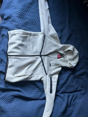 Beige nike tech  - Säljer en beige hoodie från Nike med hel dragkedja framtill och svarta detaljer längs fickor och ärm. Tröjan har luva , två sidofickor och en extra ficka på ena ärmen. Använd 2-3 gånger. Köpt från Jd. skriv för mer bilder 