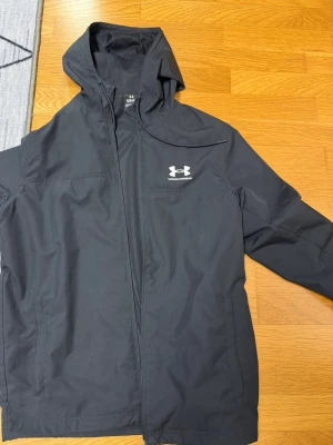 Svart vindjacka från Under Armour - Snygg svart vindjacka från Under Armour med huva och dragkedja framtill. Jackan har meshfoder på insidan och två sidofickor. Logotypen är tryckt i vitt på bröstet. Perfekt för blåsiga dagar och sportiga outfits. Finns lite smuts vid armen men det går bort. Sällan använd.