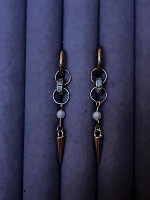Guldtonade örhängen med pärldetalj - Säljer ett par trendiga örhängen i guldtonad metall med små gnistrande stenar, vita pärldetaljer och spetsiga hängen. Örhängena har en cool kedjedesign med flera ringar och en edgy vibe. Perfekta för dig som gillar statement-smycken och vill sticka ut.