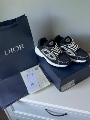 Dior b30 - Snygga svarta Dior sneakers med vita och grå detaljer samt vita skosnören. Skorna har en chunky vit sula och meshpaneler för extra komfort. Märkeslogga på plösen och sidan. Perfekta för dig som vill ha en exklusiv och trendig look.