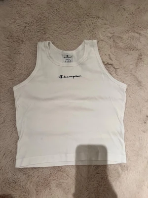 Vit linnetopp från Champion - Säljer en vit linnetopp från Champion med broderad logga framtill och liten logga bak i nacken. Toppen är ärmlös och har en klassisk, sportig look. Perfekt till sommaren och enkel att matcha med olika outfits.