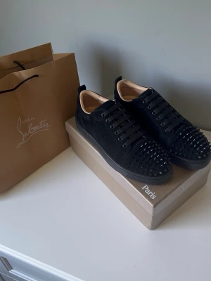 Svarta Louboutin sneakers med nitar - Säljer ett par svarta sneakers från Christian Louboutin i mocka med svarta nitar över tån. Skorna har klassisk låg siluett, svarta skosnören och en diskret logga på hälen. Insidan är beige och sulan är svart. Riktigt edgy och lyxig vibe.